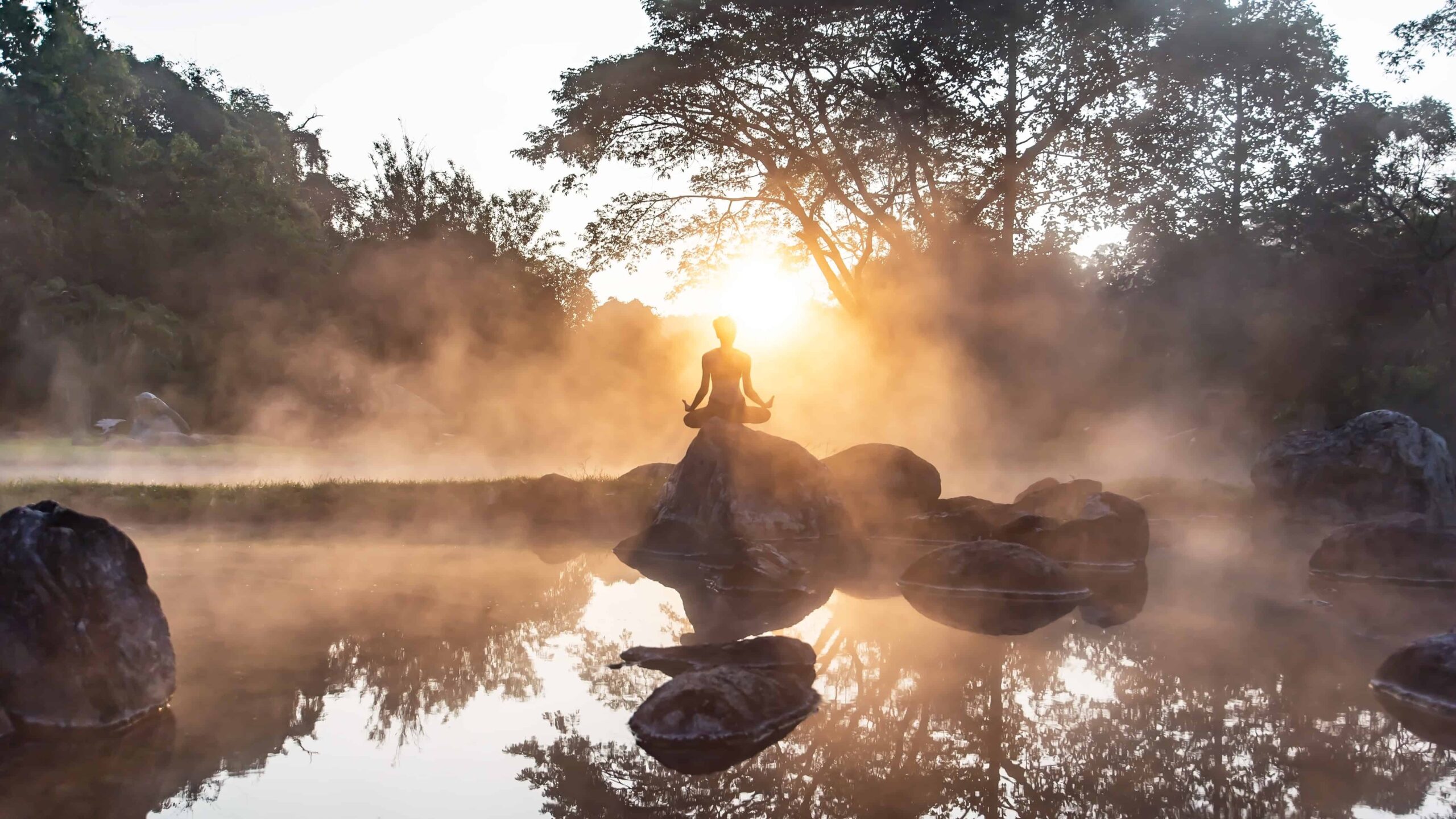 Woman sitting on a rock in a river in early morning facing the sunrise. Taking in the energy of the sun.