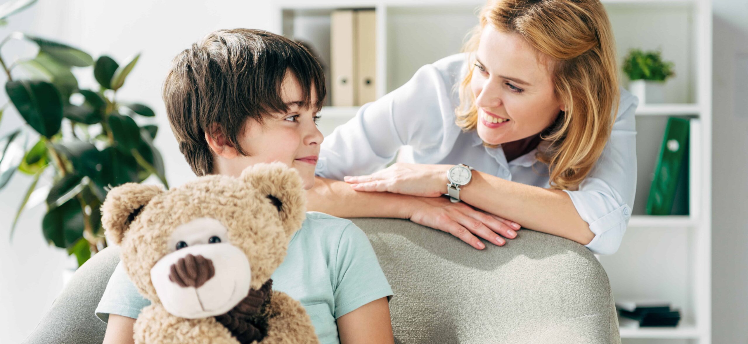 Autistic boy with a teddy on his lap, looking happy with mom smiling at him.
